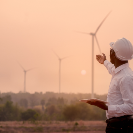 Man in hardhat outside gesturing at wind turbines during a hazy sunset