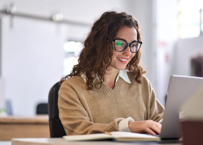 Young happy professional business woman worker employee sitting at desk working on laptop in corporate office. Smiling female student using computer technology learning online, doing web research.